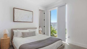 A modern bedroom with a large bed, a view of greenery through an open door, and a framed abstract painting on the wall. - Aria • Fully-Fenced Pool & Hot Tub, Luxury Design (Yucca Valley)