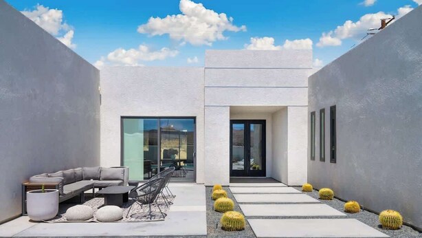 A modern outdoor patio with a gray sofa, dining table, and chairs, surrounded by white walls and large windows. - Aria • Fully-Fenced Pool & Hot Tub, Luxury Design (Yucca Valley)
