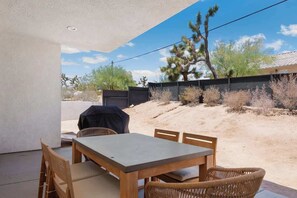 Eine Terrassengaststätte mit Tisch und Stühlen, einem Grill und Blick auf eine Wüstenlandschaft mit vereinzelter Vegetation und einem Baum.