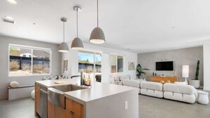 A modern kitchen with a central island, a dining area with a sofa, and a living room with a TV and plants. - Aria • Fully-Fenced Pool & Hot Tub, Luxury Design (Yucca Valley)