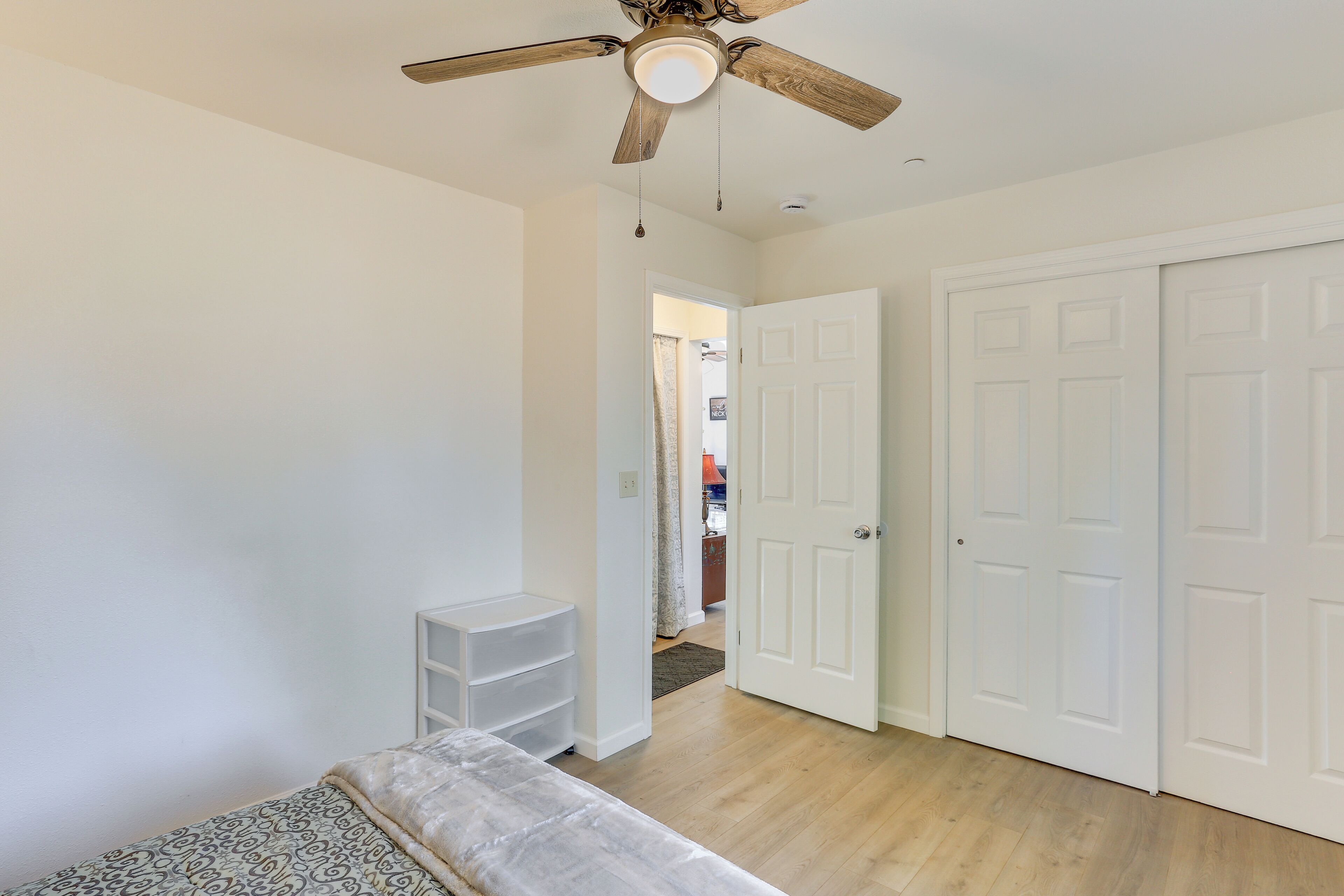 Une pièce avec un ventilateur de plafond, des murs blancs, un plancher en bois, un lit, une porte blanche et un couloir menant à une autre pièce.