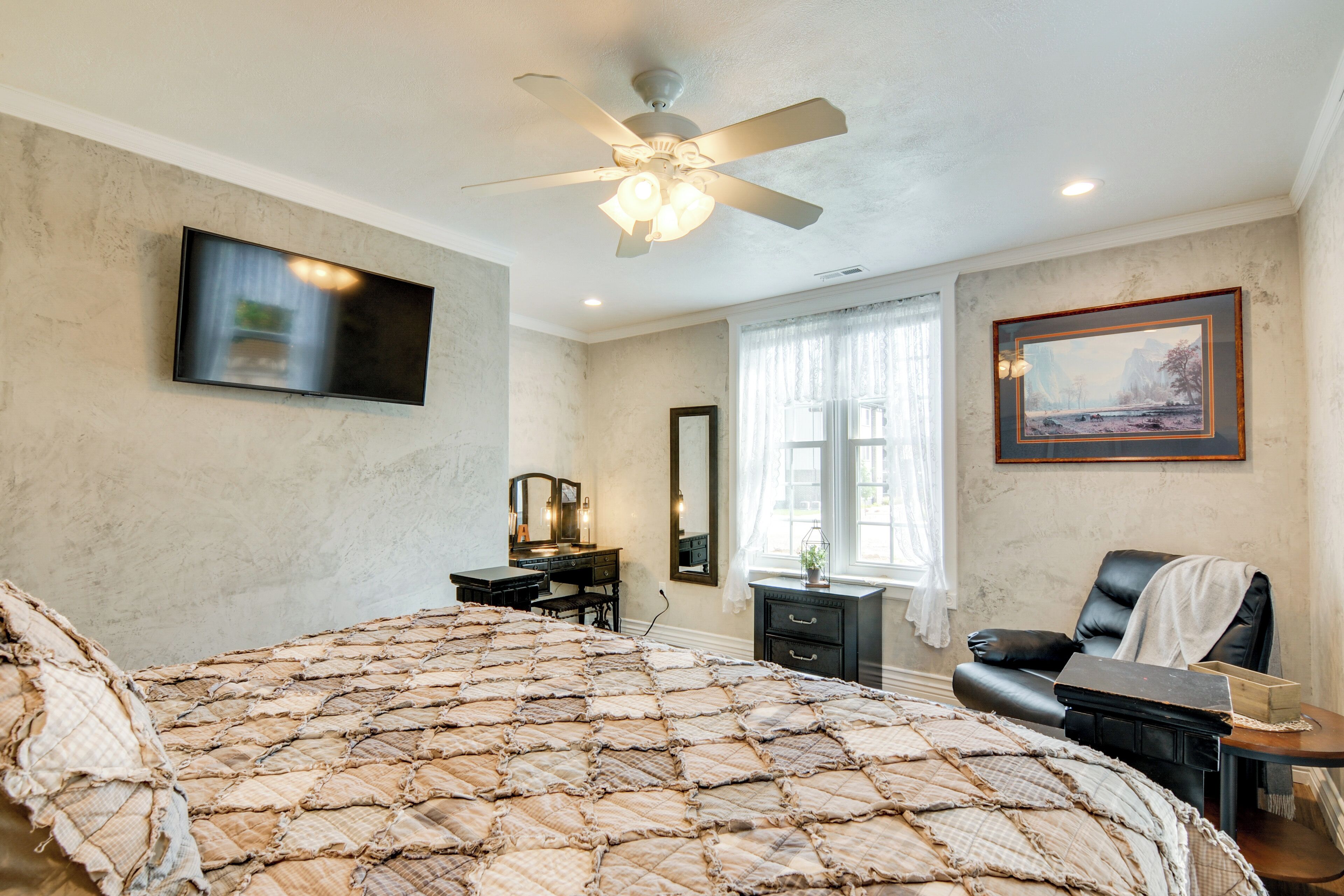 A bedroom with a large bed, a ceiling fan, a flat-screen TV, a dresser, a chair, and a window with curtains.