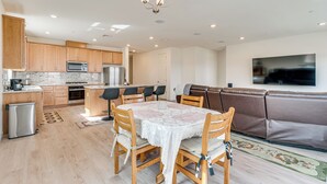 A modern kitchen with wooden cabinets, a dining table with chairs, a sofa, and a TV mounted on the wall.