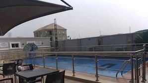 A poolside area with a table and chairs, a covered seating area, and a view of the pool and buildings in the background. - Peninsula Suites (Bwari)