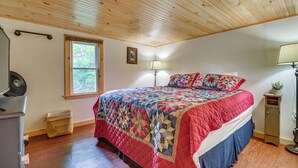 A bedroom with a wooden ceiling, a bed with a colorful quilt, a window, a lamp, and a laundry basket. - Deck + Fire Pit: Nature Lover's Retreat in Maine! (Errol)