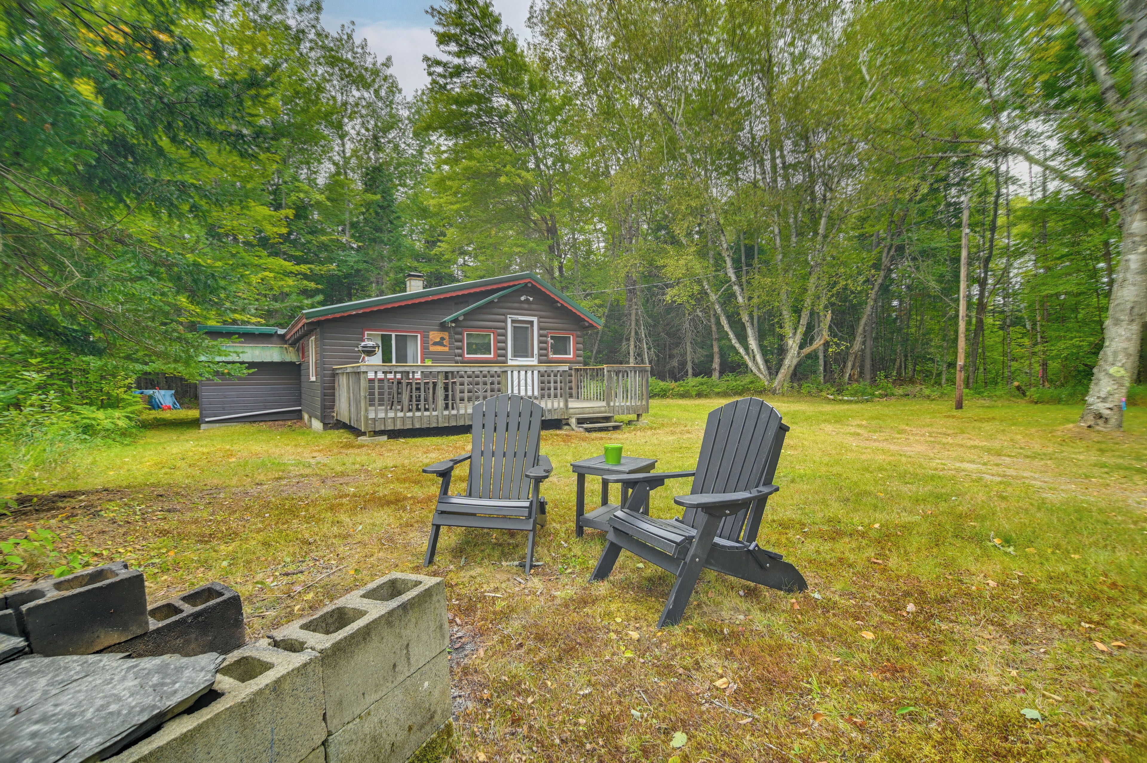 A cabin with a deck, two Adirondack chairs, and a fire pit in a wooded area.