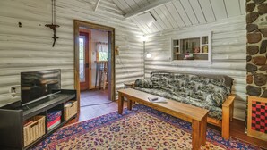 A log cabin interior with a television on a wooden stand, a patterned sofa, a wooden table, a colorful rug, and a stone accent wall. - Deck + Fire Pit: Nature Lover's Retreat in Maine! (Errol)