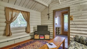 A cozy cabin interior with a wooden floor, a patterned rug, a small table with a remote control, and a bookshelf with books and baskets. - Deck + Fire Pit: Nature Lover's Retreat in Maine! (Errol)