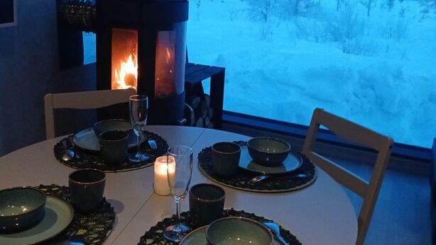 Una mesa redonda para comer con una vela, platos y cubiertos. Se observa una chimenea con una decoración de venado al fondo.