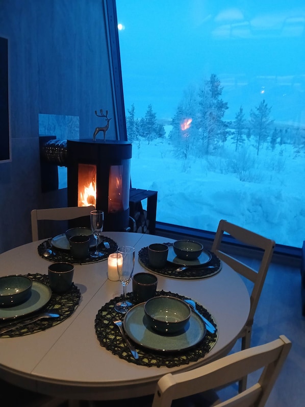 A round dining table with a candle, plates, and cutlery. A fireplace with a deer decoration is visible in the background.