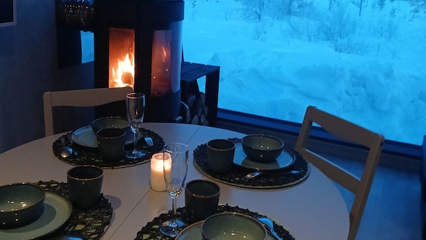A round dining table with a candle, plates, and cutlery. A fireplace with a deer decoration is visible in the background. - Lodge Arctic 4 – sauna, toit vitré & vue lac Inari (Inari)