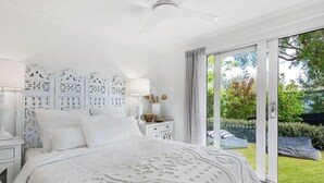 A bedroom with a large bed, a white headboard, a nightstand with a lamp, and a view of a garden through a sliding glass door. - Rye Coastal Haven : 3  BR, 2  BA House in Rye, Sleeps 6 (Rye)