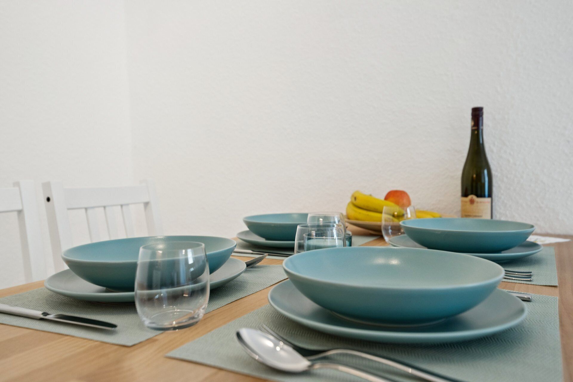 A dining table set with blue plates, glasses, and cutlery, accompanied by a bottle of wine and a bowl of fruit.