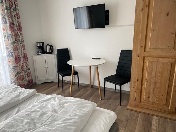 Habitación de hotel con cama, una pequeña mesa blanca, dos sillas negras, televisor montado en la pared, armario de madera y cortina estampada.
