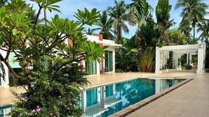 A pool area with a clear blue pool, surrounded by palm trees and a white building with green shutters. - Modern Townhouse | Condo in Sam Roi Yot (Sam Roi Yot)