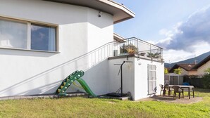 A white building with a balcony, a green slide, a basketball hoop, and a table with chairs. - Villa 'Il Gelsomino' with Mountain View, Private Terrace, EV Charging & Wi-Fi (Cuasso al Piano)