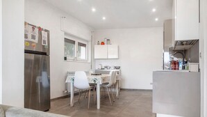 A compact kitchen with a refrigerator, dining table, chairs, and a small window. - Villa 'Il Gelsomino' with Mountain View, Private Terrace, EV Charging & Wi-Fi (Cuasso al Piano)