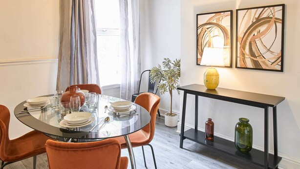 Uma sala de jantar com uma mesa de vidro redonda, cadeiras laranja e um aparador preto com vasos decorativos.