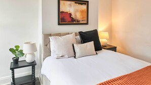 Cama bem arrumada com roupa de cama branca e um cobertor laranja. Um criado-mudo preto pequeno com uma luminária branca e uma planta em vaso fica ao lado da cama. Um quadro está pendurado na parede acima da cama.