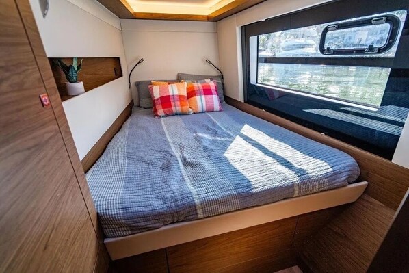 A compact bed with a checkered blanket, a small shelf with a plant, and a window with a view of the outside.