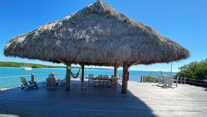 Une terrasse en bois surmontée d’une structure à toit de chaume, avec des chaises longues et des tables offrant une vue sur une étendue d’eau.