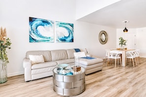 Ein Wohnzimmer mit einem beigen Sofa, einem runden Couchtisch und einem Essbereich mit weißem Tisch und Stühlen.