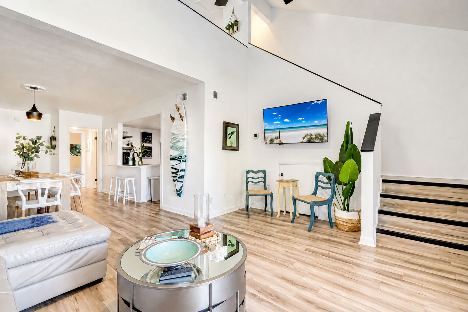 A spacious living area with a modern design, featuring a dining area, a seating area with chairs, a large window with a beach view, and a staircase.