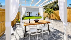 Une terrasse extérieure avec une table en bois et des chaises blanches, une pergola agrémentée de rideaux blancs, et une vue sur une pelouse verdoyante et une maison en arrière-plan.