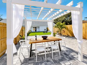 Eine Terrassengruppe im Freien mit einem Holztisch und weißen Stühlen, eine Pergola mit weißen Vorhängen sowie ein Blick auf eine grüne Rasenfläche und ein Haus im Hintergrund.