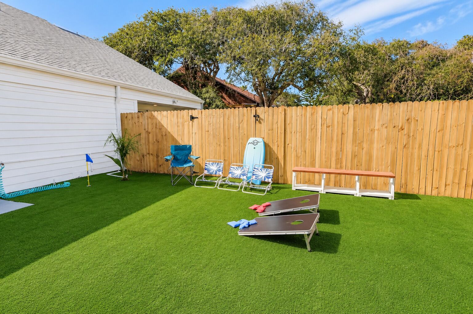 Un jardin avec une pelouse artificielle, une clôture en bois, un bâtiment blanc et du mobilier d’extérieur comprenant un banc et des chaises.