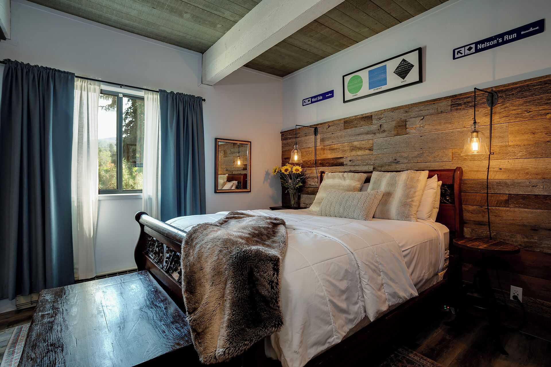 A bedroom with a wooden headboard, a bed with white bedding, a bench at the foot of the bed, a window with curtains, and a wall-mounted sign indicating 'Nelson's Run'.