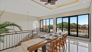 Un comedor con una larga mesa dispuesta para cuatro, un ventilador de techo, ventanas amplias y vistas al exterior.