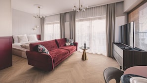 Een moderne hotelkamer met een rode bank, een bed, een televisie en grote ramen met gordijnen.
