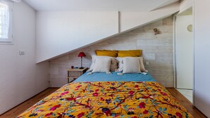 Un dormitorio con techo inclinado, una cama con colcha floral de colores, dos almohadas, una mesita de noche con lámpara y un cabecero de madera.
