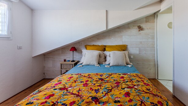 Un dormitorio con techo inclinado, una cama con colcha floral de colores, dos almohadas, una mesita de noche con lámpara y un cabecero de madera.