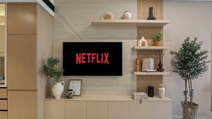 Un salon moderne avec un téléviseur fixé au mur affichant le mot « NETFLIX » en lettres rouges, des meubles en bois, une plante en pot et des objets décoratifs disposés sur des étagères.