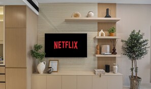 A modern living room with a mounted TV displaying "NETFLIX" in red letters, wooden cabinets, a potted plant, and decorative items on shelves. - WYZ RENT|JVC Boho Grecian Studio Pool & Gym (Dubai)