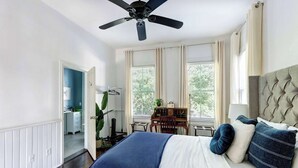 A bedroom with a bed, a ceiling fan, a desk with a chair, and a window with curtains. - The Whale Loft | Historic Charm + Balcony (Savannah)