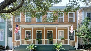 A two-story house with a front porch, American flags, and potted plants. - The Whale Loft | Historic Charm + Balcony (Savannah)