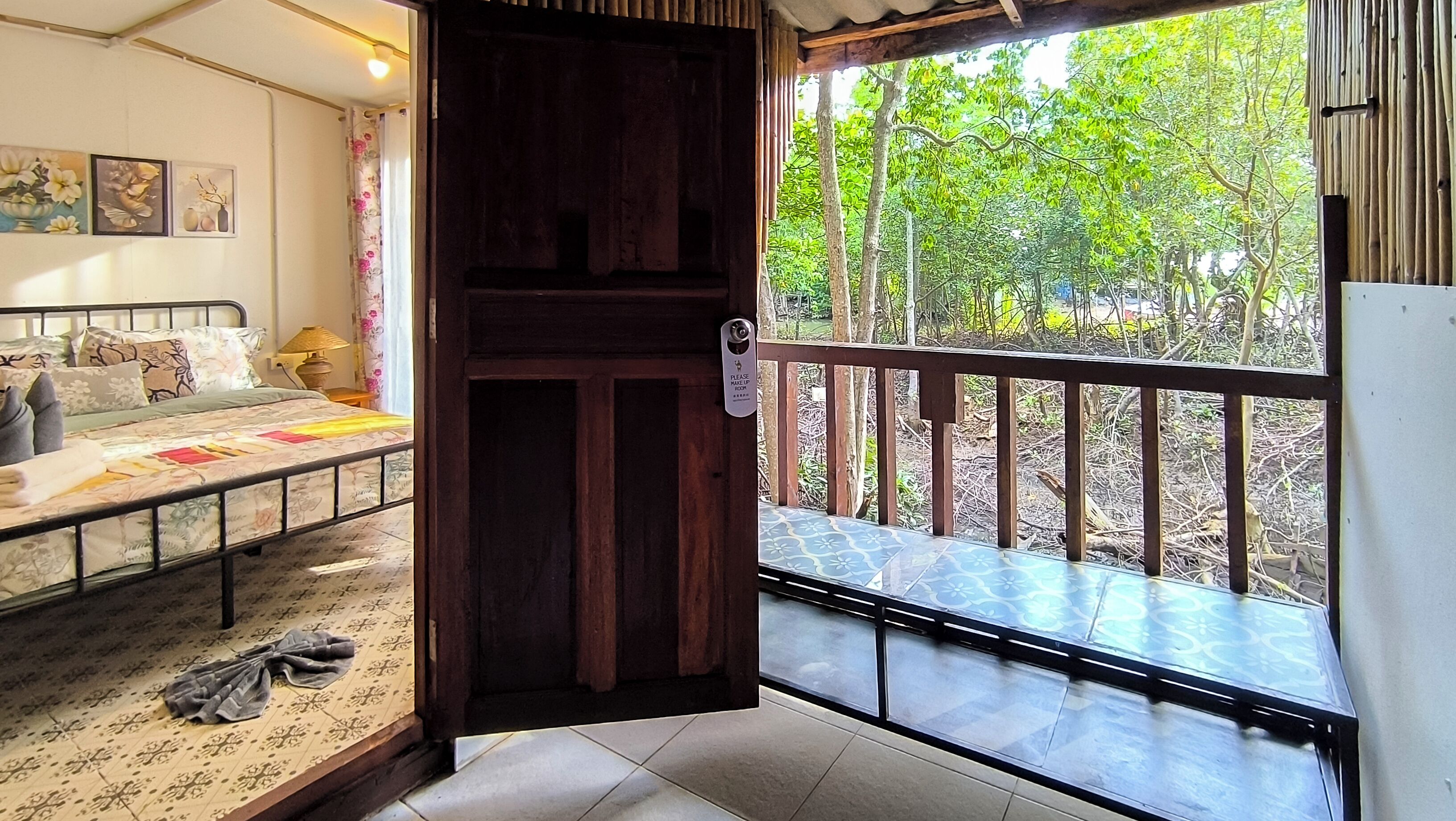A room with a bed, a small table, a chair, a window with a view of greenery, and a door with a security device.