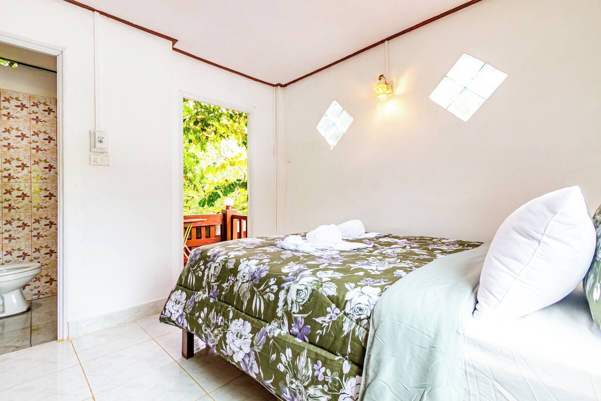 Un dormitorio con colcha estampada, inodoro blanco y ventana con vistas a la vegetación.