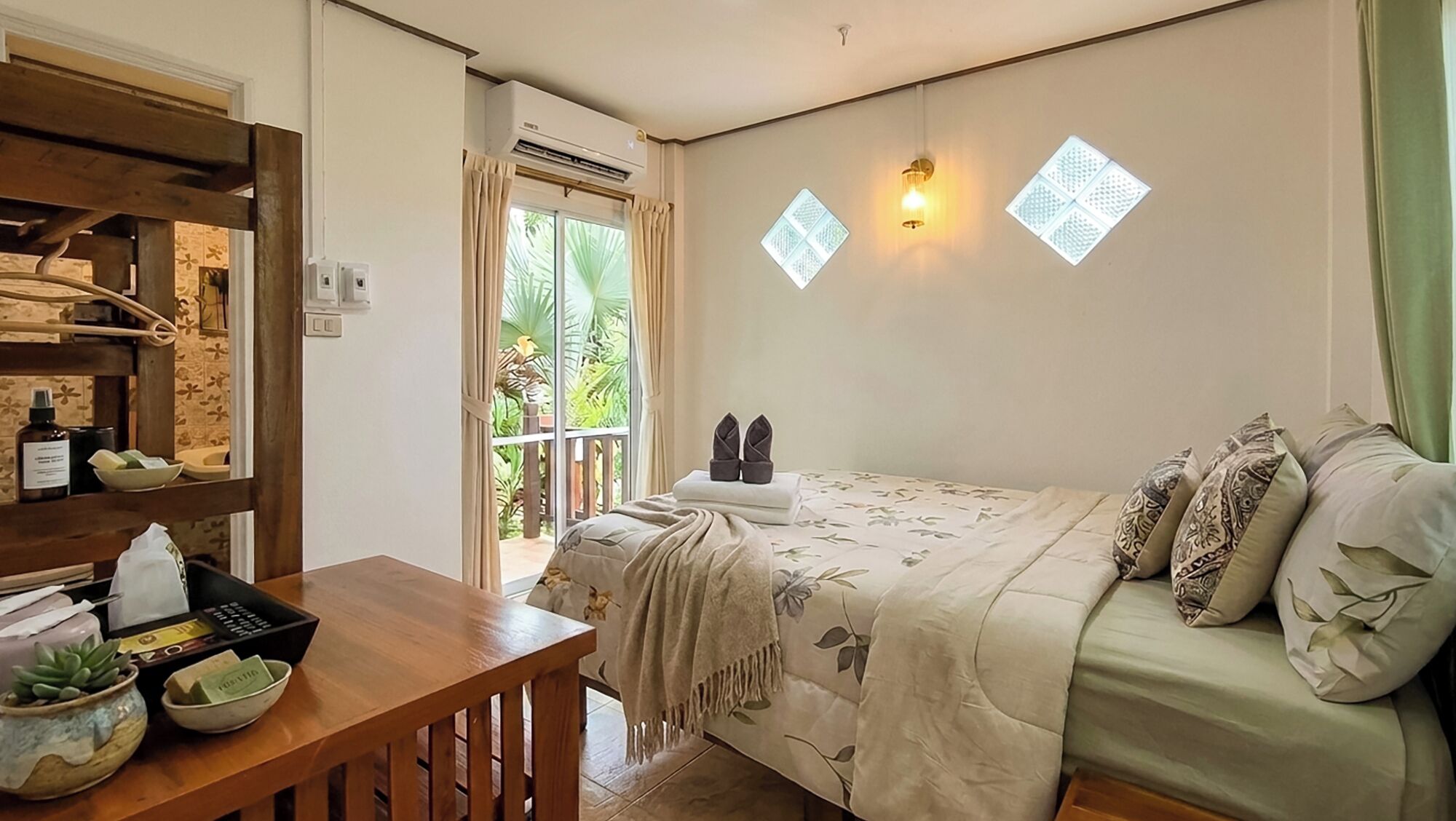 A bedroom with a bed, a wooden desk, a shelf with toiletries, and a view of greenery through a window.