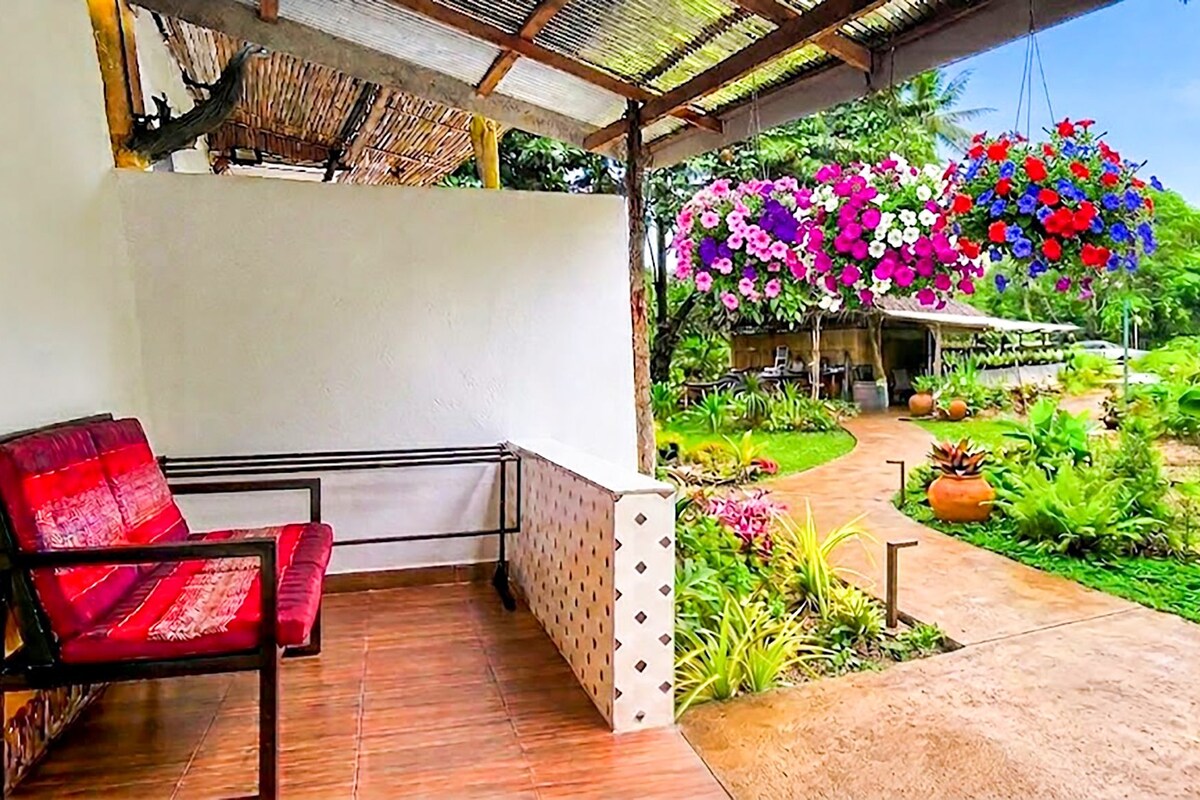Un espacio al aire libre techado con una silla roja acolchada, un sendero que conduce a un jardín con plantas variadas y cestas de flores colgantes.