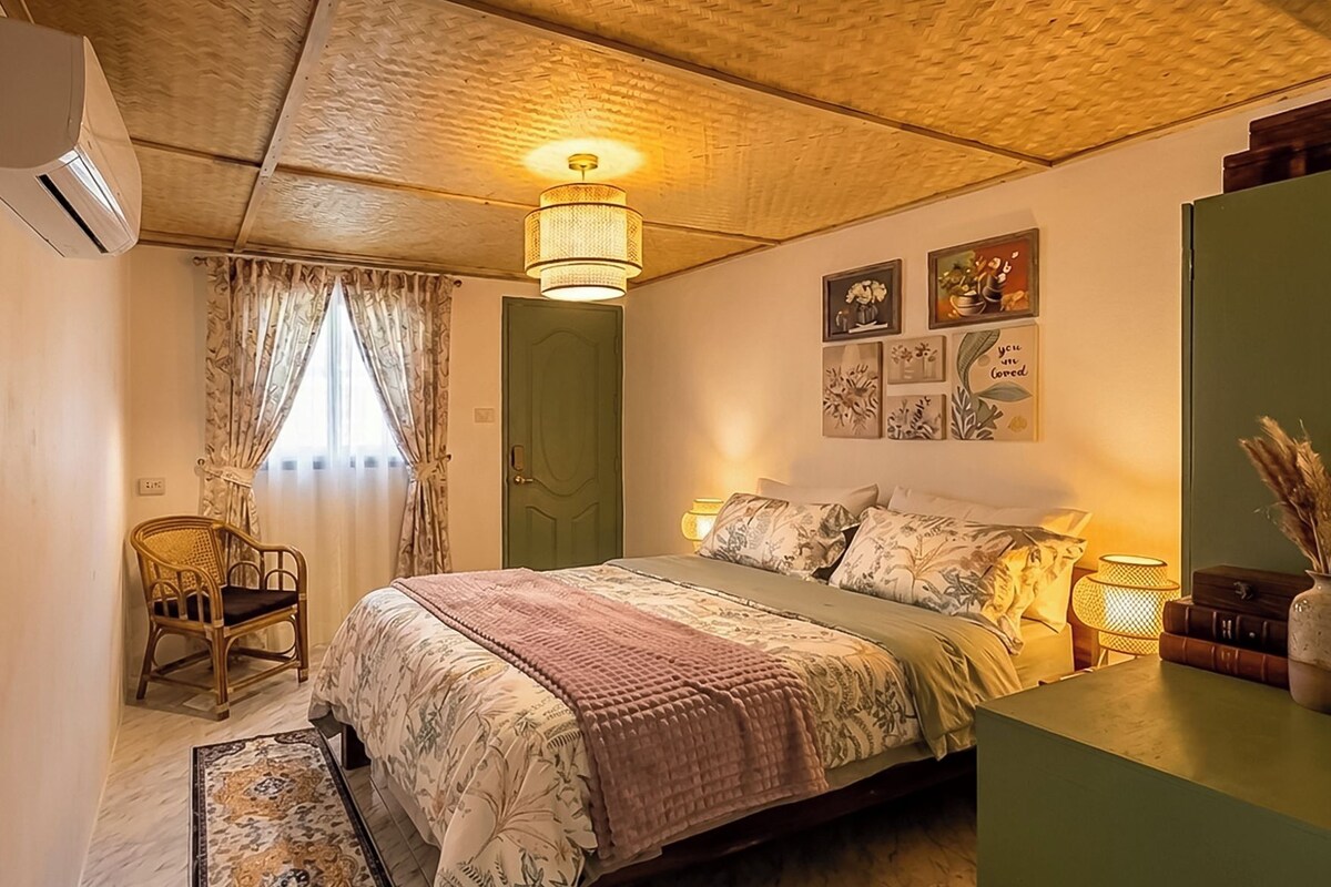 A bedroom with a bed, a chair, a bedside table, a lamp, and a wall with framed pictures.