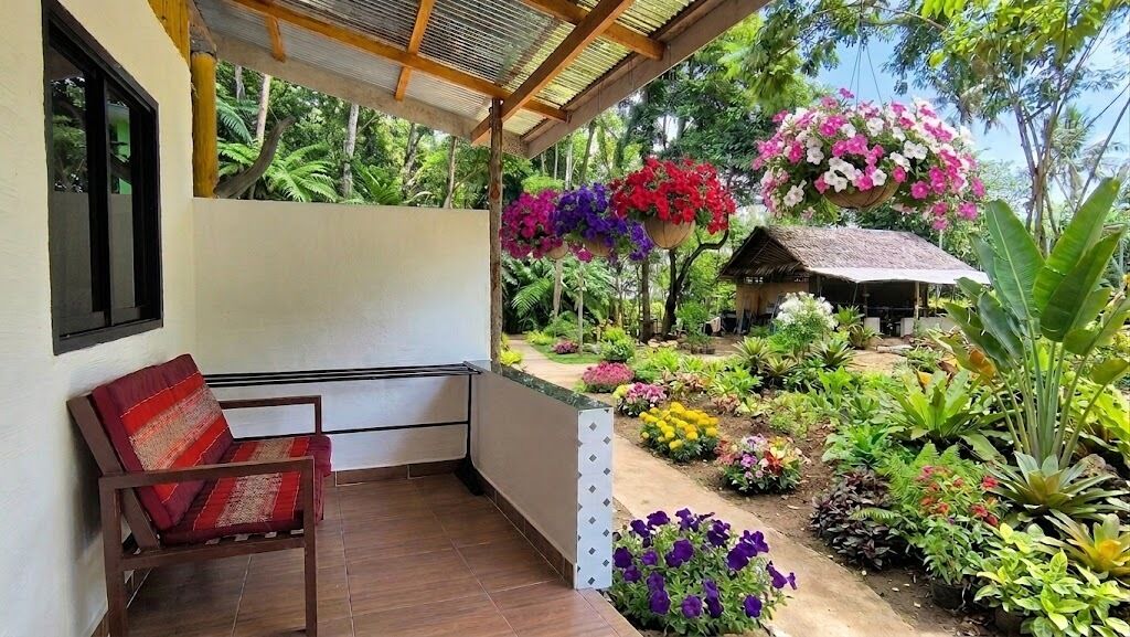 A covered outdoor area with a wooden bench, a thatched-roof structure, and a variety of potted plants and flowers.