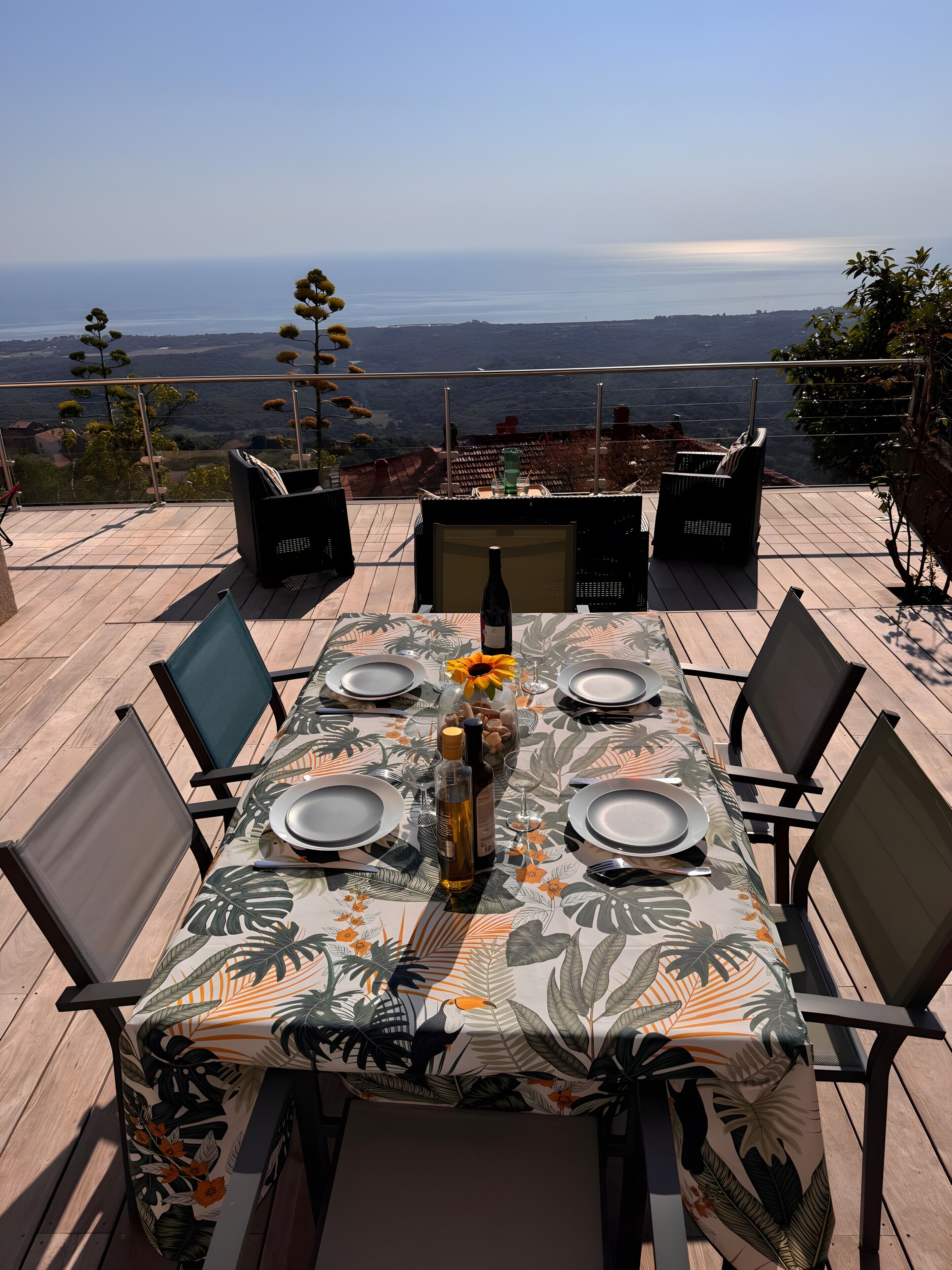 Une table à manger dressée pour six personnes, avec une nappe ornée d’un motif de feuilles tropicales, des chaises et une bouteille de vin.
