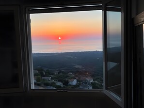 A window with a view of a sunset over a landscape with houses and hills.