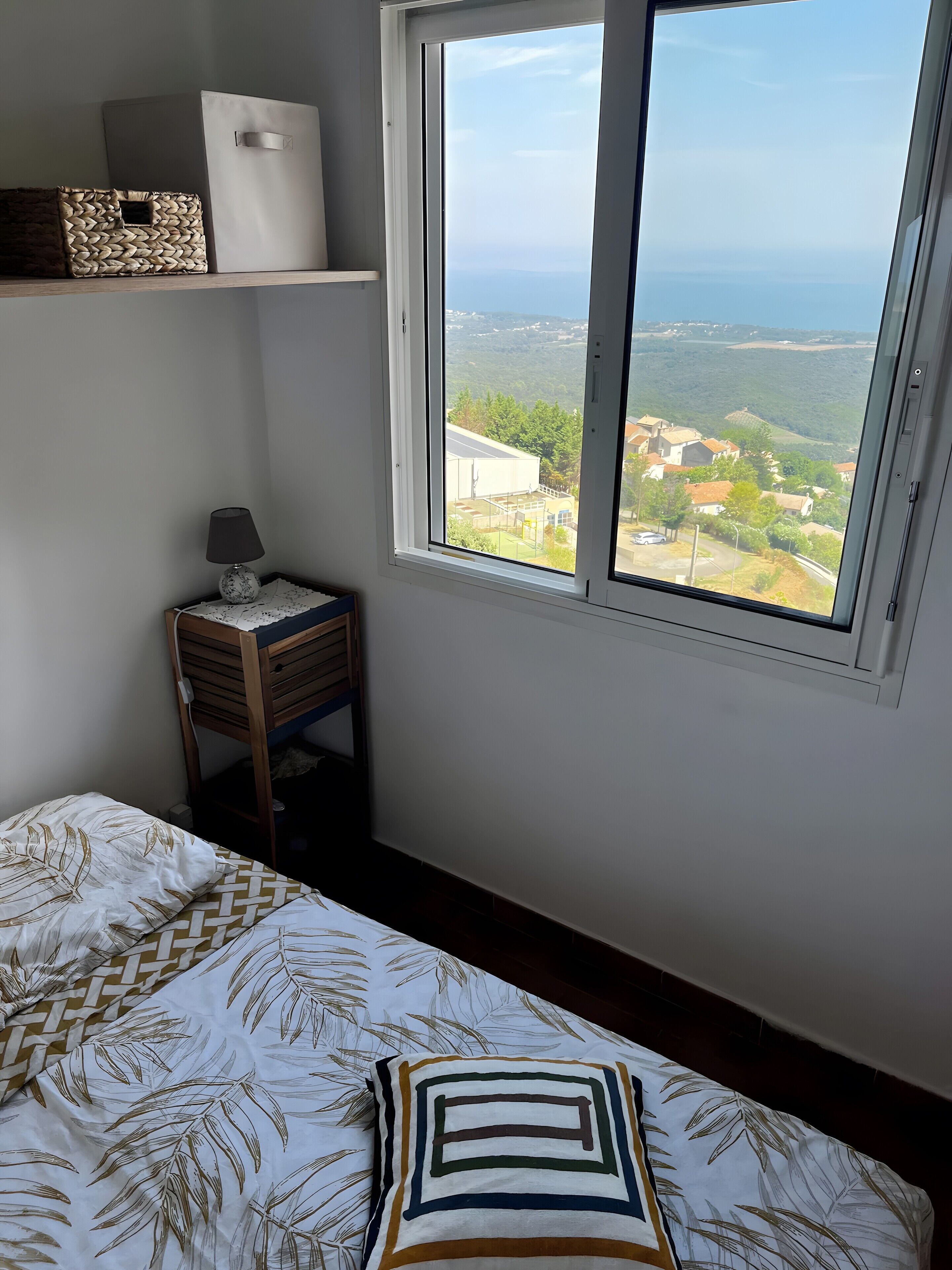 Un dormitorio con cama, mesita de noche, lámpara, ventana con vista a un paisaje y estante con objetos.