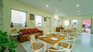 Un comedor con una mesa de madera dispuesta para cuatro personas, rodeada de sillas blancas. En primer plano, un sofá marrón con cojines rojos.