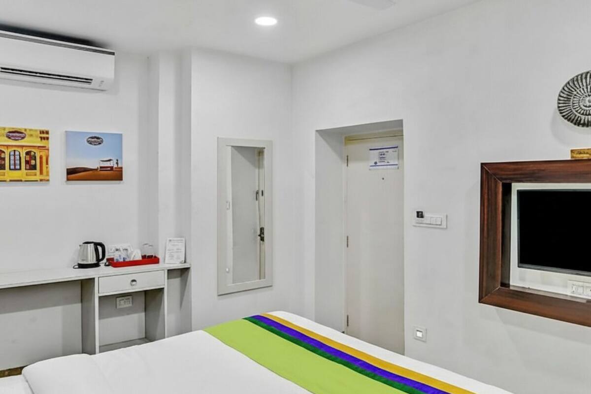 A hotel room with a bed, a desk with a kettle, a small table with a tray, a window, and a door.